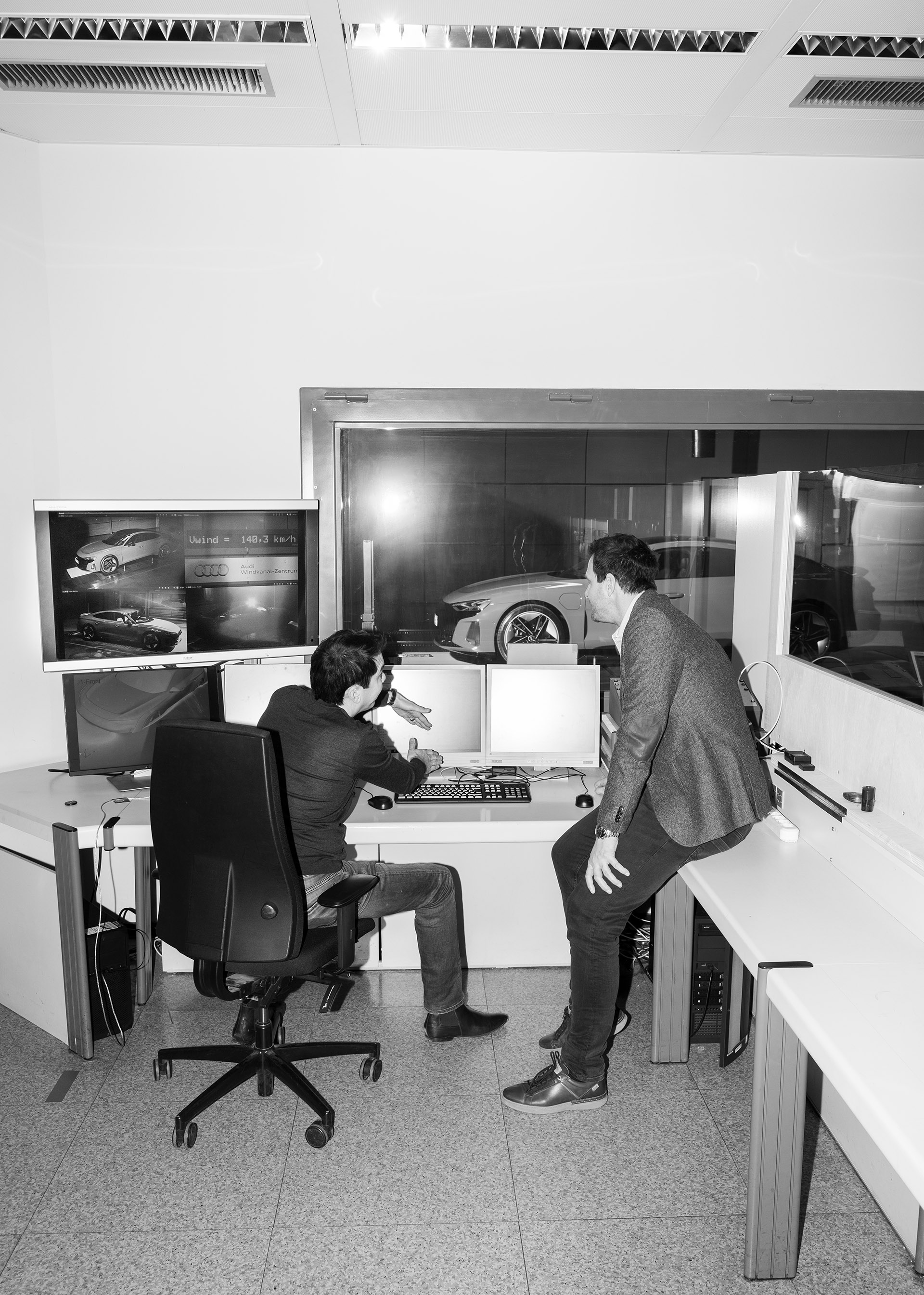 Dr. Kentaro Zens and Thomas Redenbach in discussion in front of a number of computer screens, with the Audi RS e-tron GT{ft_rs-e-tron-gt} behind a window in the background.